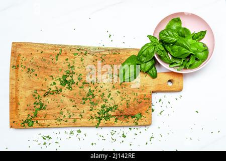 Frische Basilikumblätter in einer kleinen Schüssel neben einem hölzernen Schneidbrett auf einer weißen Oberfläche. Frisches, gesundes und natürliches Lebensmittelkonzept. Speicherplatz kopieren. Stockfoto