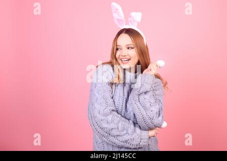 Schönes Mädchen in einem blauen Pullover auf einem rosa Hintergrund. Frau mit Hasenohren, posiert für ostern, Mädchen lächelt, Platz für Text. Stockfoto