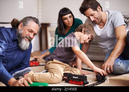 Familienspaß auf der Eisenbahn. Eine kleine Aufnahme eines kleinen Kindes, das mit einem Zugfahrsatz spielt, während seine Familie zuschaut. Stockfoto