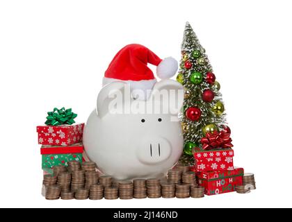 Weißes Schweinebank aus Porzellan mit Weihnachtsmütze, umgeben von Geldstapeln, Quartieren, Weihnachtsbaum und Geschenken. Isoliert auf Weiß Stockfoto