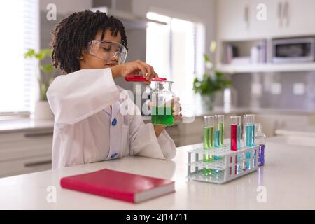 Hispanischer Junge mit Labormantel und Brille, der chemische Lösung aus dem Reagenzglas in den Becher gießt Stockfoto