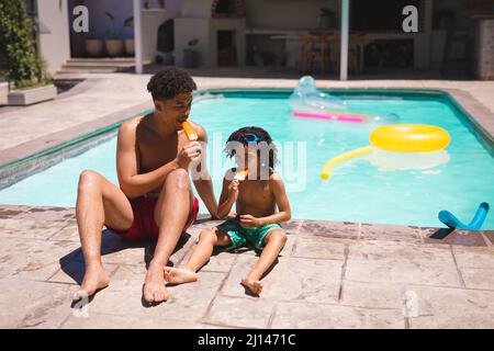 Hispanischer Vater und Sohn essen Eisdips, während sie am sonnigen Tag zusammen am Pool sitzen Stockfoto