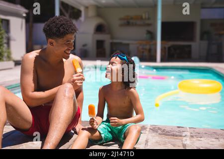 Fröhlicher hispanischer Vater und Sohn, der an einem sonnigen Tag zusammen am Pool Eis isst Stockfoto