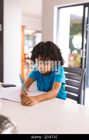 Netter hispanischer Junge, der beim Schreiben im Buch am Esstisch sitzt Stockfoto