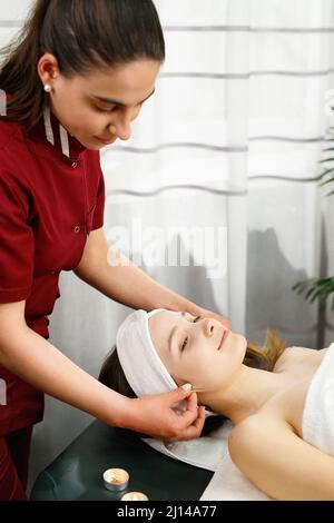 Ganzkörperfoto einer Masseurin in roter Uniform, die einer Kundin die Ohren massiert, die im Spa-Salon massiert wird. Stockfoto