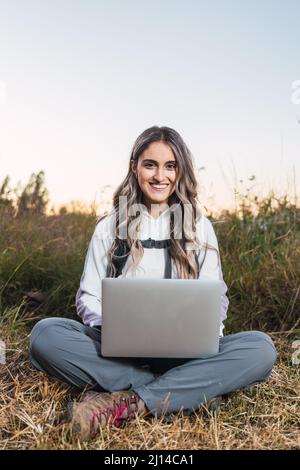 Junge kaukasische Frau mit Rucksack, die mitten auf dem Feld einen Laptop für die Telearbeit benutzt. Digitaler Nomade. Stockfoto