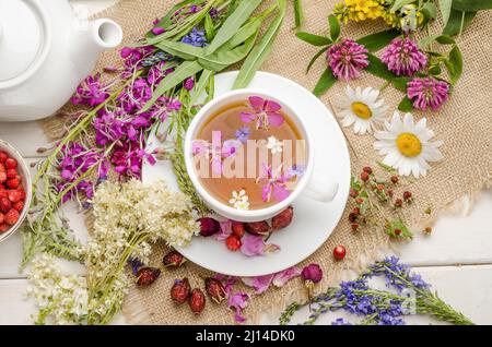 Kräutertee mit Hagebutte, Kamille und Mädesüß in einer weißen Tasse auf einem weißen Holztisch mit Blumen, flach liegend Stockfoto