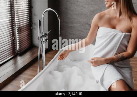 Junge Dame in Handtuch gewickelt Vorbereitung zum Baden, sitzen in der Badewanne und berühren Wasser aus dem Wasserhahn, Ernte Stockfoto