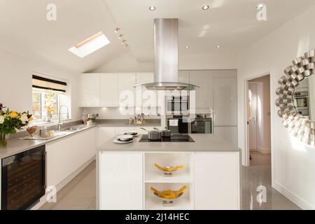 Weiße Küche im modernen Haus, Inselzentrale mit Herd und Dunstabzugshaube, Hochglanz-Einheiten, Fliesenboden. Stockfoto