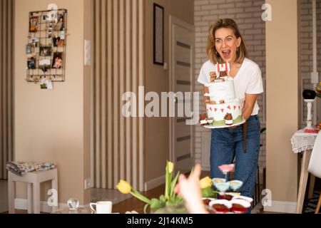 Frau steht und hält einen großen Geburtstagskuchen. Mutter bringt Essen auf den Tisch, einen weißen Kuchen zum Geburtstag eines Kindes. Stockfoto