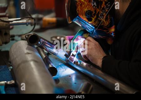 Arbeiter in einem Helm mit Flammen schweißen den Stahlteil eines Motorrads mit einem Argon-Schweißgerät und Handschuhen in einer Garage Stockfoto