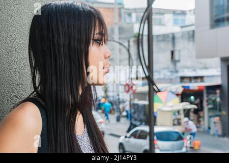 Schöne braune Latina-Frau, die neben einer Betonwand steht und auf eine andere Person wartet, während sie die Leute auf der Straße vorbeisieht. Konzept Stockfoto