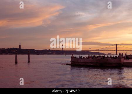 Touristen, die den Sonnenuntergang am Pier von Cais das Colunas am Ufer des Tejo in Lissabon genießen, mit der Ponte de 25 Abril im Hintergrund, Portugal. Stockfoto