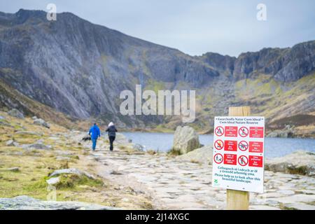 Rückansicht eines Paares, das am See entlang in den walisischen Bergen des Snowdonia National Park läuft; Schild im Vordergrund, das die Öffentlichkeit über die Beschränkungen des Naturreservats informiert. Stockfoto