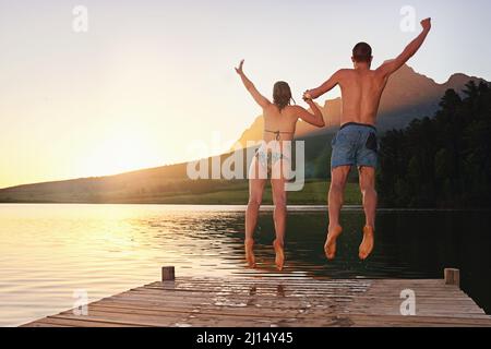 Auf dem Sprung. Rückansicht eines jungen Paares in Badeanzügen, das bei Sonnenuntergang von einem Dock springt. Stockfoto