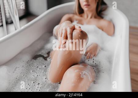 Verfahren zur Sauberkeit und häuslichen Schönheit. Junge Dame, die sich in der Badewanne entspannt und Schaum und berührende Beine genießt Stockfoto