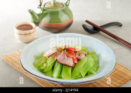 Gebratener chinesischer Salat mit Sichuan-geräucherten Bacon-Bits in einem Gericht, isoliert auf einer Holzmatte, Seitenansicht auf grauem Hintergrund Stockfoto