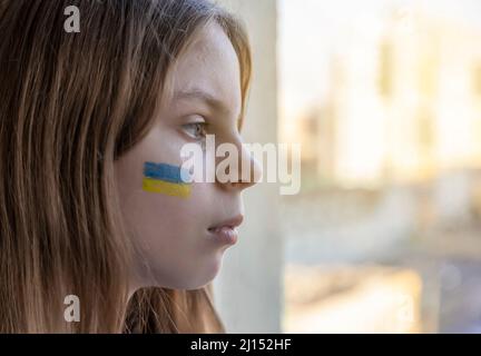 Krieg. Russlands Invasion der Ukraine. Porträt eines Kindes mit Tränen in den Augen, das auf dem Tisch sitzt und aus dem Fenster schaut. Das Blau und Gelb Stockfoto