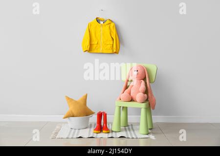 Stuhl mit Spielzeug, Gummistiefeln und Kinderregenmantel an der hellen Wand Stockfoto