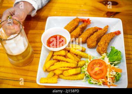 Auf dem Tisch Eine Platte mit Garnelen in Teig, pommes frites und Gemüsesalat. Ein Mann hält ein Glas Bier in der Hand. Abendessen mit Meeresfrüchten. Stockfoto