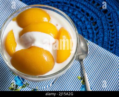 Pfirsich-Scheiben in Sirup mit Joghurt von oben auf einem blau gestreiften Handtuch fotografiert und ein weiterer Teil aus blauem Häkeln und einem Löffel auf der Seite, diese elem Stockfoto