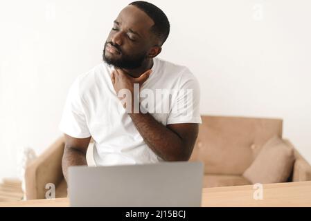 Ein schwarzer Mann arbeitet von zu Hause aus mit einem Laptop und leidet unter Halsschmerzen. Ein afroamerikanischer Mann beginnt krank zu werden und eine Halsschmerzen zu spüren Stockfoto