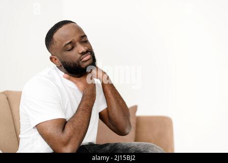 afroamerikanischer Mann, der unter Nackenschmerzen leidet, während er zu Hause auf dem Sofa im Wohnzimmer Muskelschmerzen massiert. Neurologisches Gesundheitsproblem Stockfoto