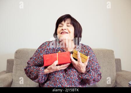 Eine Oma hält zu Hause ein rotes und ein gelbes Geschenk auf ihren Händen auf ihrem Sofa Stockfoto