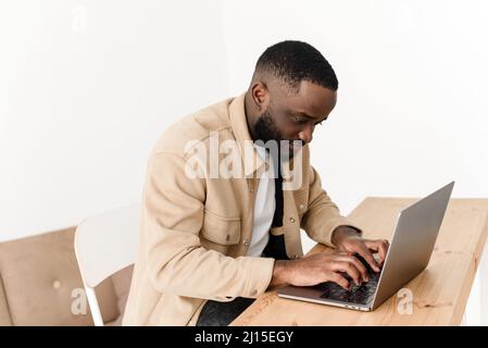 Fokussierter afroamerikanischer Mann, der von zu Hause aus am Laptop arbeitet. Der freie Programmierer arbeitet am Tisch Stockfoto