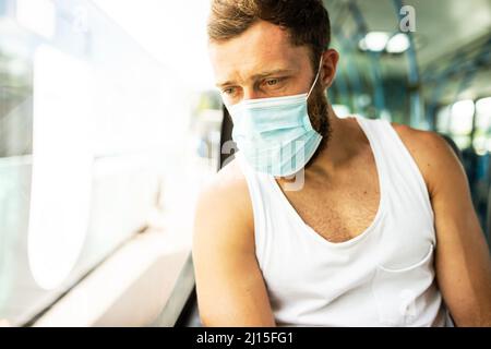 Ein gutaussehender Kerl in einem Unterhemd, der mit öffentlichen Verkehrsmitteln mit einer Schutzmaske auf seinem Gesicht reist, respektiert epidemiologische Maßnahmen Stockfoto