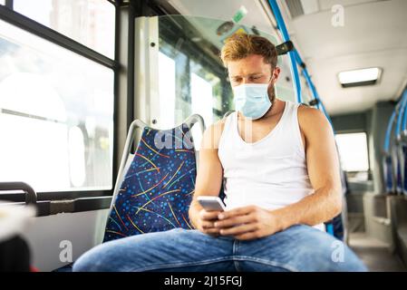 Ein gutaussehender Kerl in einem Unterhemd, der mit öffentlichen Verkehrsmitteln mit einer Schutzmaske auf seinem Gesicht reist, respektiert epidemiologische Maßnahmen und überprüft Stockfoto