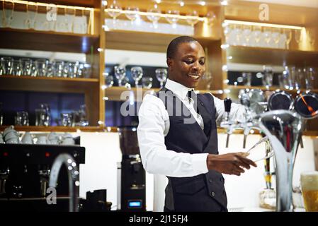 Ich zeige allen meinen Kunden eine gute Zeit. Zugeschnittene Aufnahme eines gut gekleideten Barkeepers, der hinter der Theke steht. Stockfoto