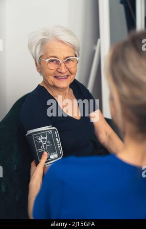Zufriedene und fröhliche ältere Rentnerin, die ihren Druck von einem professionellen Gesundheitshelfer messen ließ. Vertikale Aufnahme. Hochwertige Fotos Stockfoto