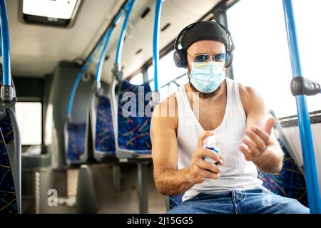 Hübscher blonder Kerl im Unterhemd, der mit öffentlichen Verkehrsmitteln mit Schutzmaske, Sonnenbrille und Kopfhörern reist, respektiert epidemiologische Maßnahmen und Stockfoto