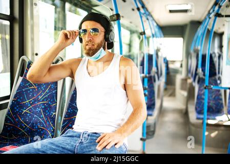 Ein gutaussehender blonder Kerl in einem Unterhemd, der mit einer Schutzmaske, Sonnenbrille und Kopfhörern mit öffentlichen Verkehrsmitteln reist, respektiert epidemiologische Fragen Stockfoto