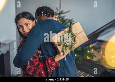Interrassisches Paar feiert Weihnachten. Umarmung zwischen zwei Ehepartnern. Kaukasische Frau, die der Kamera ein Geschenk zeigt. Hochwertige Fotos Stockfoto