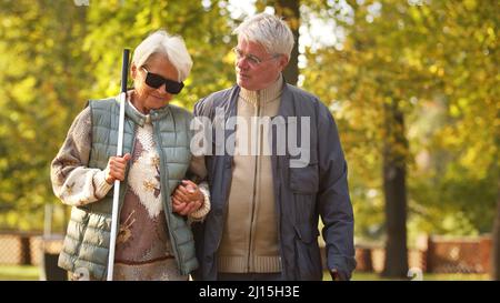 Älterer kaukasischer Mann geht mit seiner blinden Frau in einer Brille und hält sich im Park auf Behinderte Blinde unterstützen Konzept Medium shot copy space. Hochwertige Fotos Stockfoto