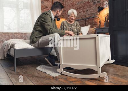 kaukasischer eleganter Novize Papa und seine ältere Mutter sitzen neben der weißen Wiege, wo Enkelkind nippt. Hochwertige Fotos Stockfoto