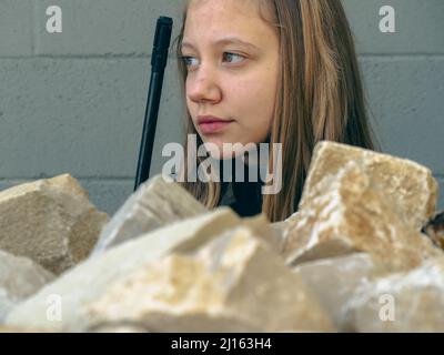 kaukasisch blondes jugendliches Soldatenmädchen an einem Kontrollpunkt mit einem Gewehr und einer camogrünen Militäruniform während eines Konflikts in einem Kriegsgebiet Stockfoto