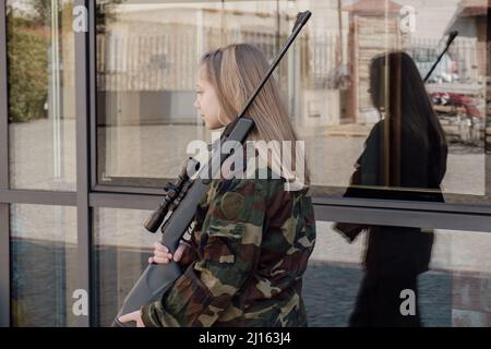 kaukasisch blondes jugendliches Soldatenmädchen an einem Kontrollpunkt mit einem Gewehr und einer camogrünen Militäruniform während eines Konflikts in einem Kriegsgebiet Stockfoto