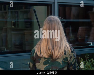 kaukasisch blondes jugendliches Soldatenmädchen an einem Kontrollpunkt mit einem Gewehr und einer camogrünen Militäruniform während eines Konflikts in einem Kriegsgebiet Stockfoto