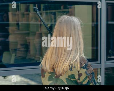 kaukasisch blondes jugendliches Soldatenmädchen an einem Kontrollpunkt mit einem Gewehr und einer camogrünen Militäruniform während eines Konflikts in einem Kriegsgebiet Stockfoto