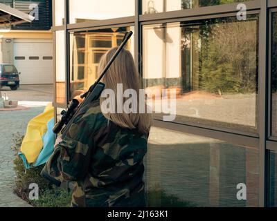 kaukasisch blondes jugendliches Soldatenmädchen an einem Kontrollpunkt mit einem Gewehr und einer camogrünen Militäruniform während eines Konflikts in einem Kriegsgebiet Stockfoto