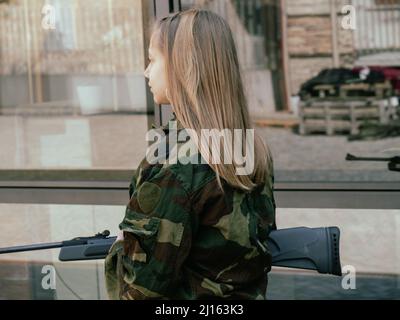 kaukasisch blondes jugendliches Soldatenmädchen an einem Kontrollpunkt mit einem Gewehr und einer camogrünen Militäruniform während eines Konflikts in einem Kriegsgebiet Stockfoto