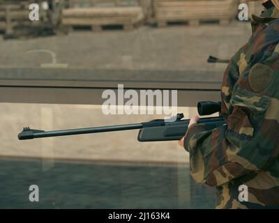 kaukasisch blondes jugendliches Soldatenmädchen an einem Kontrollpunkt mit einem Gewehr und einer camogrünen Militäruniform während eines Konflikts in einem Kriegsgebiet Stockfoto