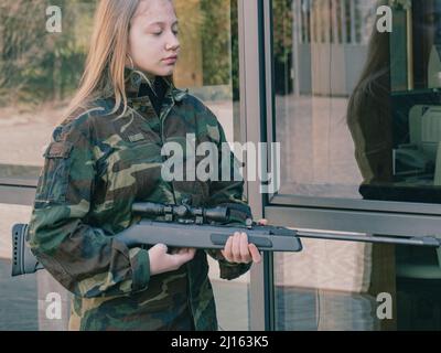 kaukasisch blondes jugendliches Soldatenmädchen an einem Kontrollpunkt mit einem Gewehr und einer camogrünen Militäruniform während eines Konflikts in einem Kriegsgebiet Stockfoto