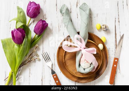 Ostertisch mit bemalten Eiern, lila Tulpe und Besteck auf festlichem Tisch. Tischeinstellung für Happy Easter Day. Draufsicht flach liegend. Stockfoto