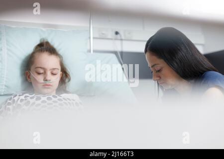 Besorgte Eltern, die im Krankenhauszimmer sitzen, während sie sich im Krankenhaus ausruhen, krankes Mädchen. Schlafende kranke kleine Tochter, die sich nach einem medizinischen Eingriff im Klinikbett ausruhte, während die traurige Mutter neben ihr wartete. Stockfoto