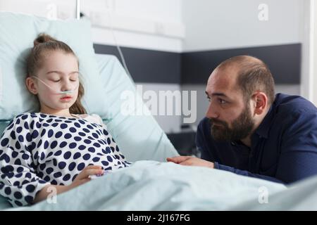 Nachdenklicher trauriger Vater, der die schlafende kranke kleine Tochter ansieht, während er im Kinderkrankenhaus-Patientenzimmer ist. Krankes kleines Mädchen, das sich nach einem medizinischen Eingriff ausruhte, während der verstörte Vater neben ihr saß. Stockfoto