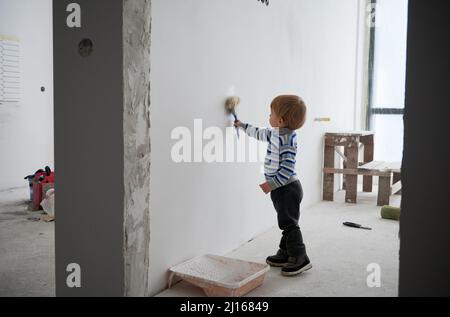 Seitenansicht des kleinen Kindes Zeichnung Wand speziellen Pinsel für die Reparatur in weißer Farbe zu Hause. Konzept der Prozesslackierung Wand im Zimmer mit guter Stimmung zu Hause während der Renovierung Wohnung. Stockfoto